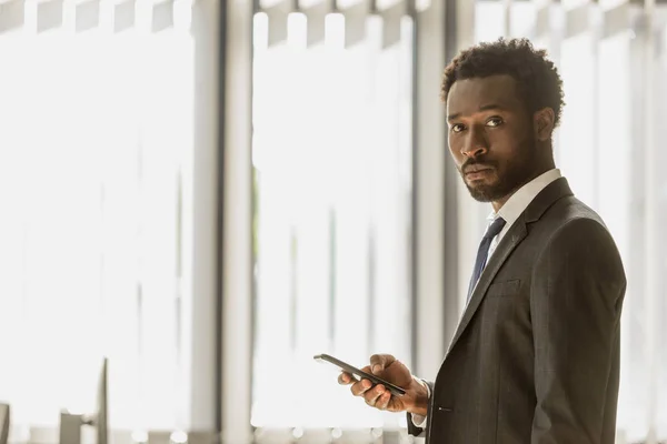 Hombre de negocios afroamericano serio sosteniendo teléfono inteligente y mirando a la cámara - foto de stock