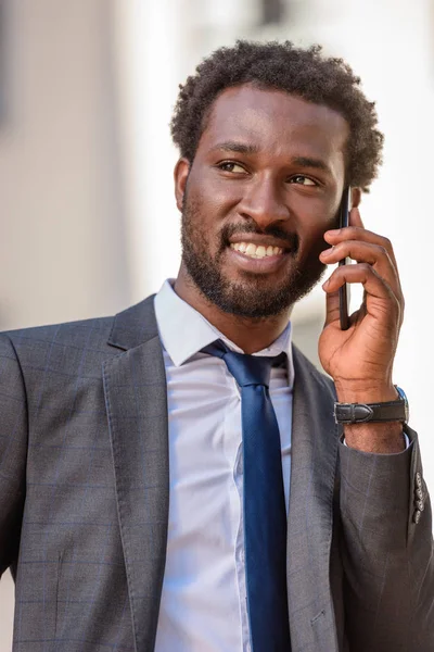 Guapo afroamericano hombre de negocios hablando en smartphone y sonriendo mientras mira hacia otro lado - foto de stock