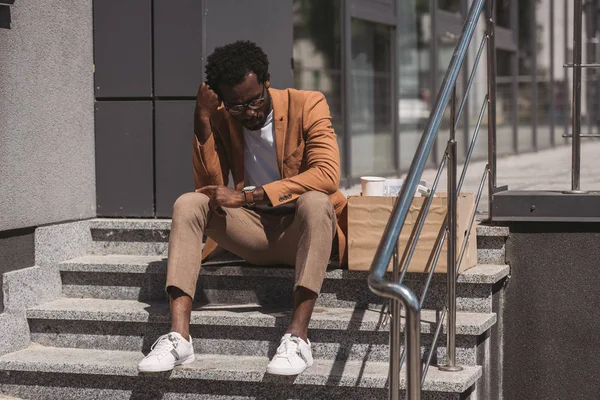 Bouleversé homme d'affaires afro-américain assis sur les escaliers près de la boîte en carton et tenant la main près de la tête — Photo de stock