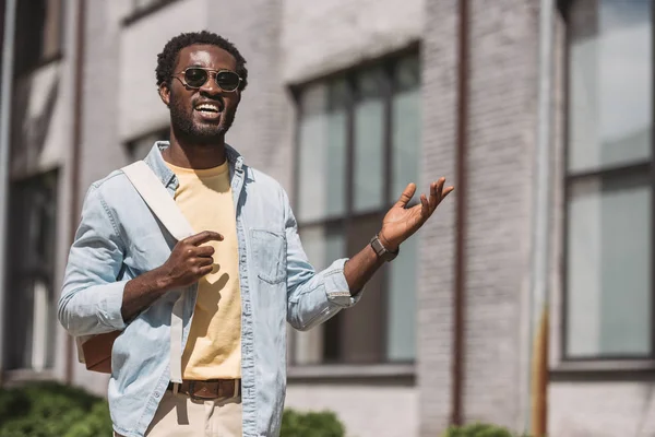 Alegre afroamericano hombre en gafas de sol sonriendo y haciendo gestos mientras mira a la cámara - foto de stock