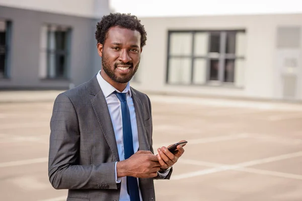 Guapo hombre de negocios afroamericano sosteniendo teléfono inteligente, mirando a la cámara y sonriendo - foto de stock