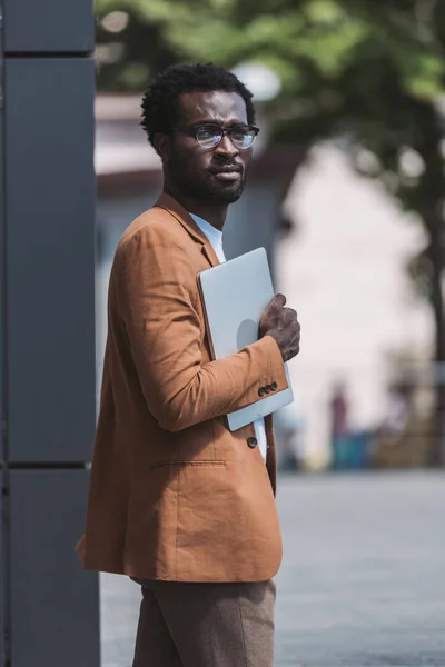 Pensativo afroamericano hombre de negocios mirando la cámara mientras sostiene el ordenador portátil - foto de stock