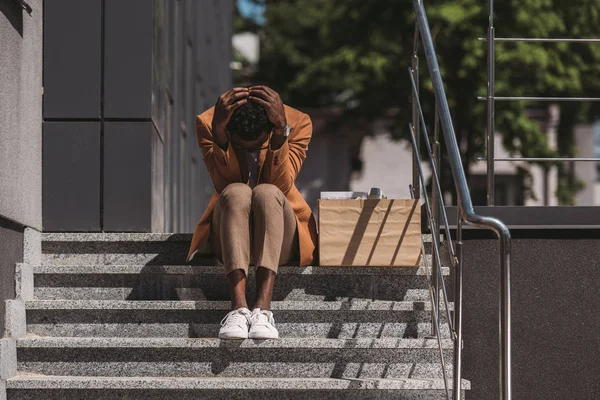 Uomo d'affari africano americano depresso seduto sulle scale vicino a scatola di cartone e tenendosi per mano sulla testa — Foto stock