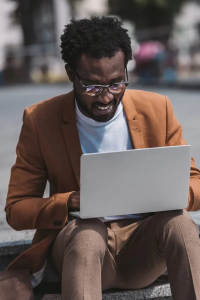 African american businessman in glasses using laptop while sitting on parapet — Stock Photo