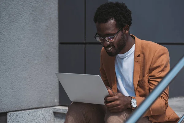 Bel homme d'affaires afro-américain utilisant un ordinateur portable tout en étant assis sur les escaliers — Photo de stock