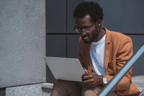 Bonito Africano americano empresário usando laptop enquanto sentado em escadas — Fotografia de Stock