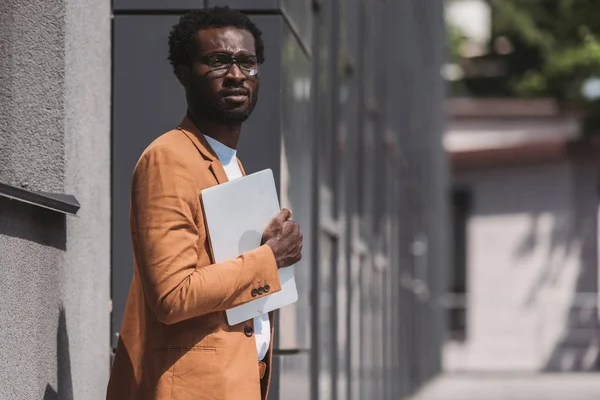 Thoughtful african american businessman holding laptop and looking away on street — Stock Photo