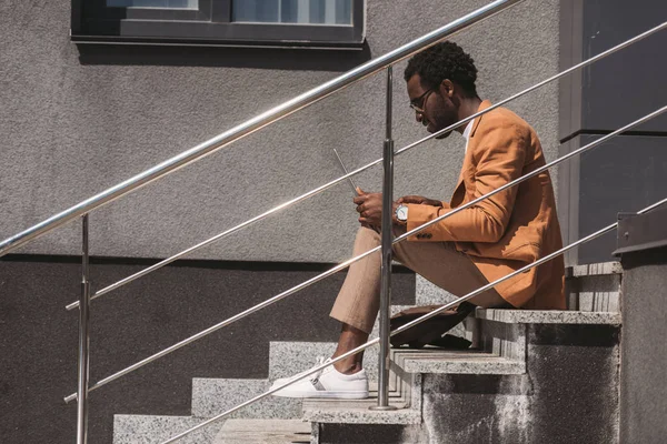 Vista lateral do homem de negócios americano africano elegante sentado nas escadas e usando laptop — Fotografia de Stock