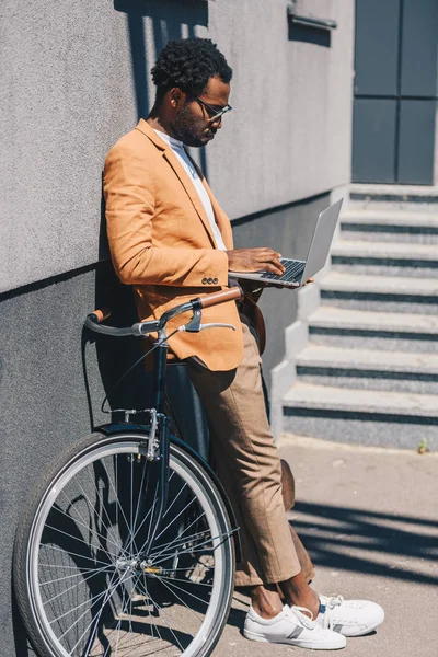 Elegante hombre de negocios afroamericano de pie cerca de la bicicleta y el uso de la computadora portátil - foto de stock
