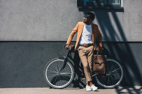 Apuesto hombre de negocios afroamericano de pie junto a la pared cerca de la bicicleta y mirando hacia otro lado - foto de stock