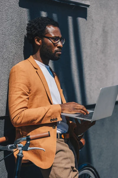 Homme d'affaires afro-américain réfléchi dans des lunettes en utilisant un ordinateur portable tout en se tenant debout avec vélo près du mur — Photo de stock