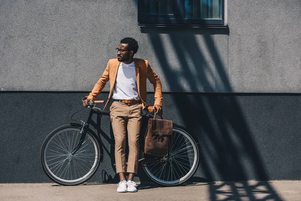 Apuesto hombre de negocios afroamericano mirando hacia otro lado mientras está de pie cerca de la bicicleta - foto de stock