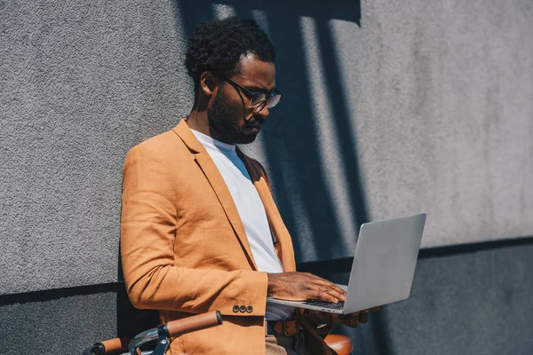 Concentrated african american businessman using laptop while standing by grey wall — Stock Photo