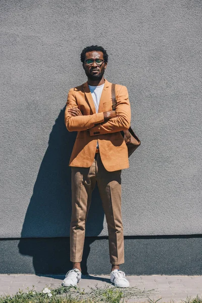 Guapo, elegante hombre de negocios afroamericano de pie con los brazos cruzados por la pared en la calle - foto de stock