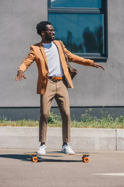 Guapo, elegante hombre de negocios afroamericano montando longboard en la calle - foto de stock