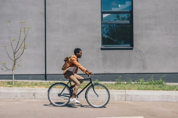 Elegante hombre de negocios afroamericano con mochila montar en bicicleta en la calle soleada - foto de stock