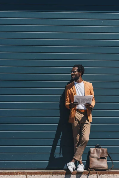 Hombre de negocios afroamericano elegante mirando hacia otro lado mientras que está parado por la pared y el periódico de celebración - foto de stock