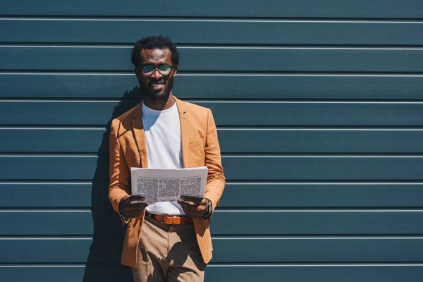 Bel homme d'affaires afro-américain tenant le journal et regardant la caméra tout en se tenant par le mur sur la rue — Photo de stock