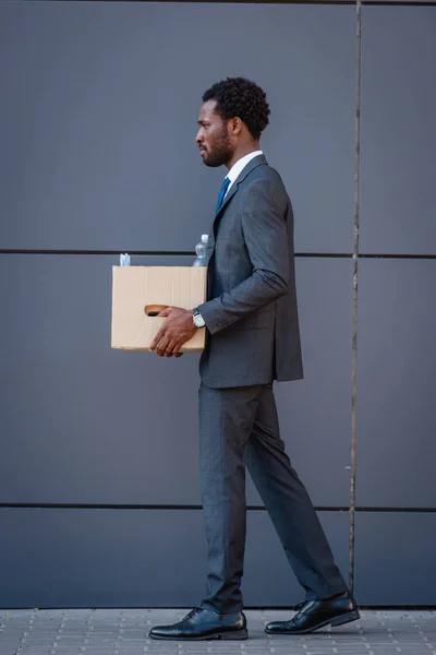 Side view of fired african american businessman holding carton box while walking on street — Stock Photo