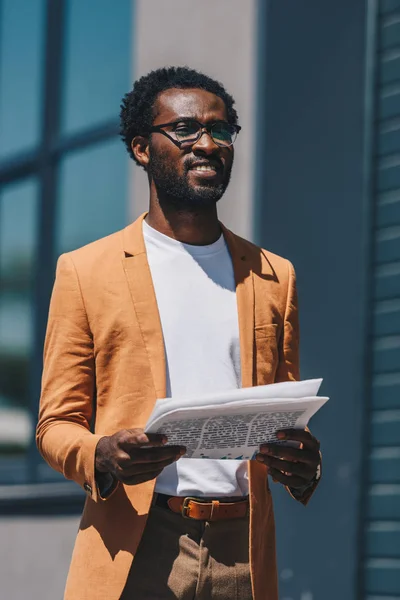 Guapo afroamericano hombre de negocios en gafas sosteniendo periódico y mirando hacia otro lado - foto de stock