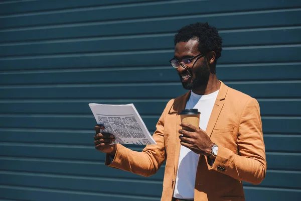 Allegro uomo d'affari afroamericano che legge il giornale mentre tiene in mano il caffè per andare — Foto stock