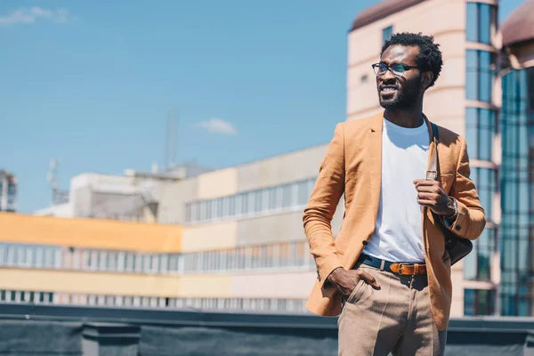 Bonito, elegante empresário afro-americano de mãos dadas e olhando para longe com edifícios em segundo plano — Fotografia de Stock