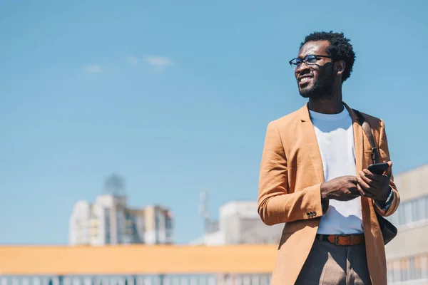 Alegre hombre de negocios afroamericano utilizando teléfono inteligente y sonriendo con el cielo azul en el fondo - foto de stock