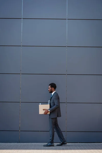 Side view of fired african american businessman carrying carton box while walking along wall — Stock Photo