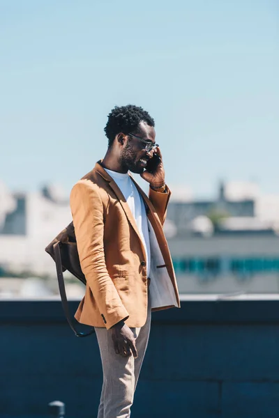 Homem de negócios americano africano elegante falando no smartphone e sorrindo no telhado — Fotografia de Stock