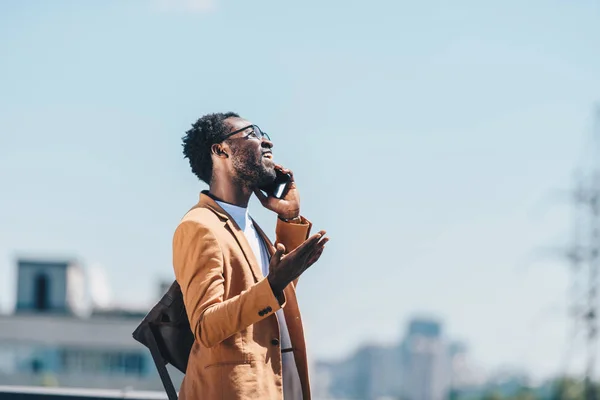 Sonriente hombre de negocios afroamericano hablando en smartphone y haciendo gestos con el cielo azul en el fondo - foto de stock