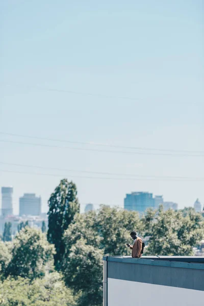 Афроамериканський чоловік використовує смартфон, стоячи на даху з блакитним небом на фоні — стокове фото