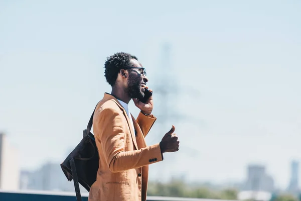 Fröhlich afrikanisch-amerikanischer Geschäftsmann spricht auf dem Smartphone und zeigt den Daumen nach oben beim Gehen auf dem Dach — Stockfoto