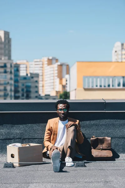 Пригнічений афроамериканець бізнесмен сидить на даху біля картонної коробці — стокове фото