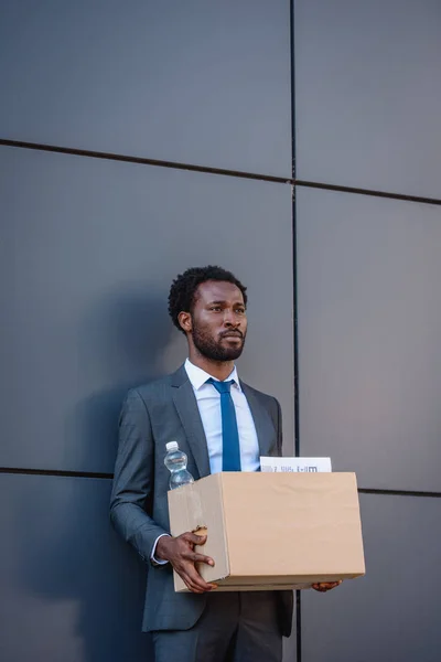 Dismissed, upset african american businessman holding carton box and looking away while standing near wall — Stock Photo