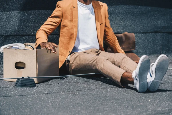 Cropped view of dismissed african american businessman sitting on rooftop near carton box — Stock Photo