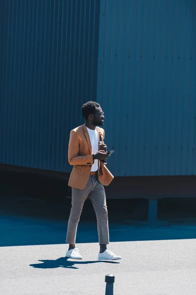 Elegante hombre de negocios afroamericano sosteniendo el teléfono inteligente y mirando hacia otro lado mientras camina en la azotea - foto de stock