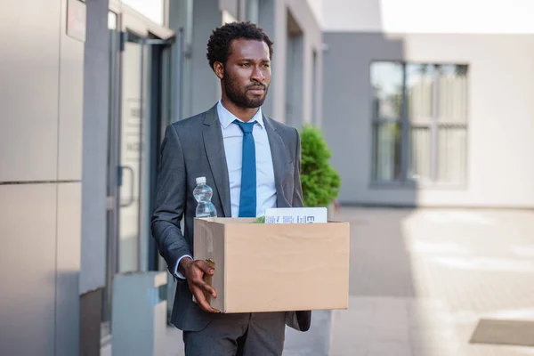 Fired, upset african american businessman looking away while holding cardboard box — Stock Photo