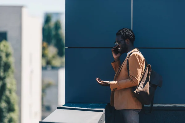 Улыбающийся африканский американский бизнесмен разговаривает на смартфоне и жестикулирует стоя на крыше — стоковое фото