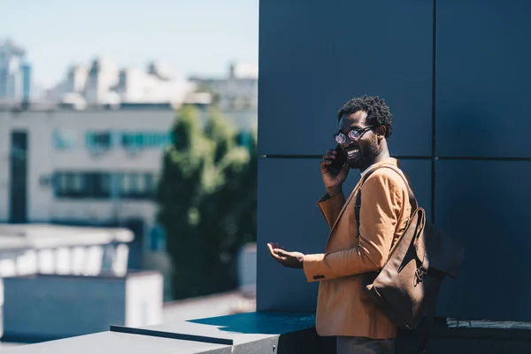 Alegre hombre de negocios afroamericano hablando en teléfono inteligente y sonriendo mientras está de pie en la azotea - foto de stock