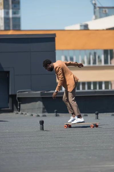 Stylish african american businessman riding longboard on rooftop — Stock Photo