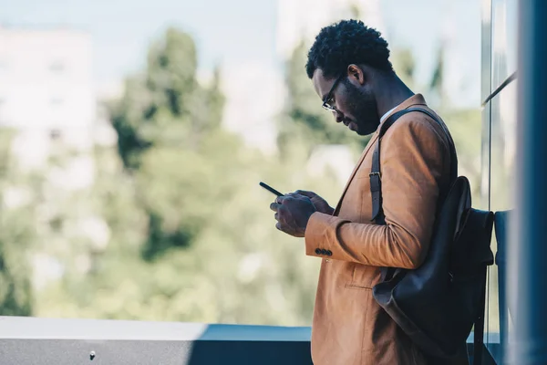 Hombre de negocios afroamericano serio de pie en la azotea y utilizando el teléfono inteligente - foto de stock