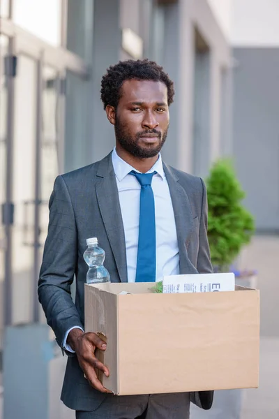 Congédié, contrarié homme d'affaires afro-américain tenant boîte en carton et regardant la caméra — Photo de stock