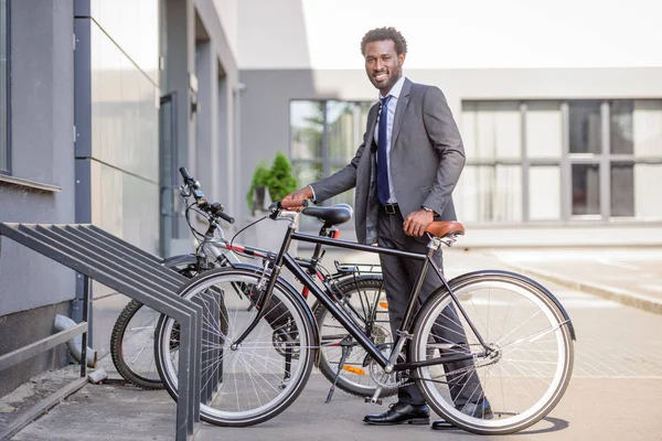Guapo afroamericano hombre de negocios sonriendo a la cámara mientras toma bicicleta desde el estacionamiento - foto de stock