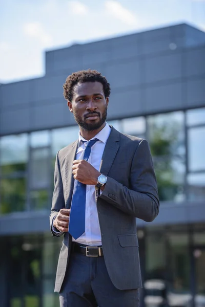 Confiado hombre de negocios afroamericano tocando corbata y mirando hacia otro lado - foto de stock