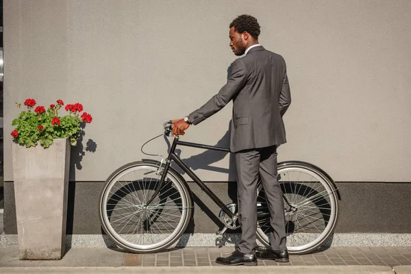 Uomo d'affari afroamericano in giacca e cravatta con bicicletta vicino al muro e fiore rosso in vaso da fiori — Foto stock