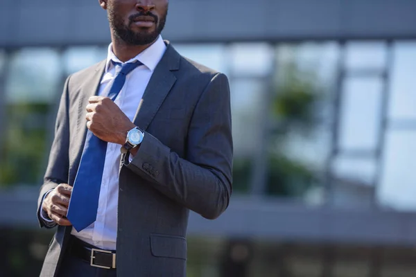 Vista parcial del empresario afroamericano tocando corbata mientras está parado en la calle - foto de stock