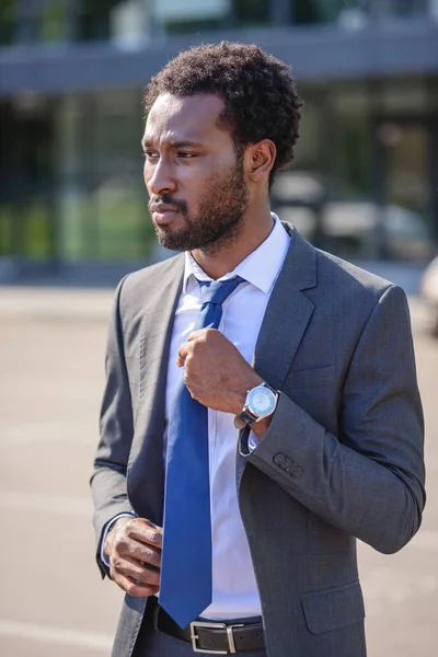 Hombre de negocios afroamericano serio tocando corbata y mirando hacia otro lado en la calle - foto de stock