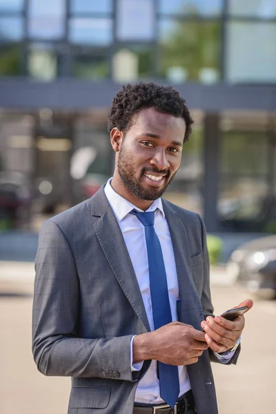 Guapo, sonriente hombre de negocios afroamericano mirando a la cámara mientras usa el teléfono inteligente - foto de stock