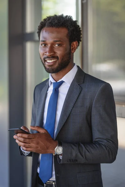 Guapo, alegre hombre de negocios afroamericano sonriendo a la cámara mientras usa el teléfono inteligente - foto de stock