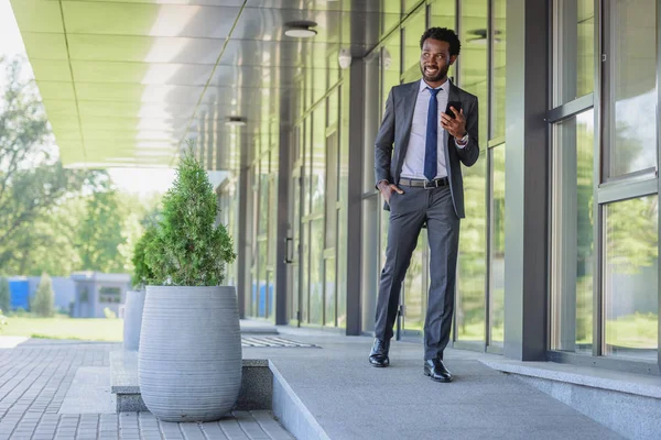 Fröhlich afrikanisch-amerikanischer Geschäftsmann hält Smartphone in der Hand, während er an Bürogebäuden entlangläuft — Stockfoto